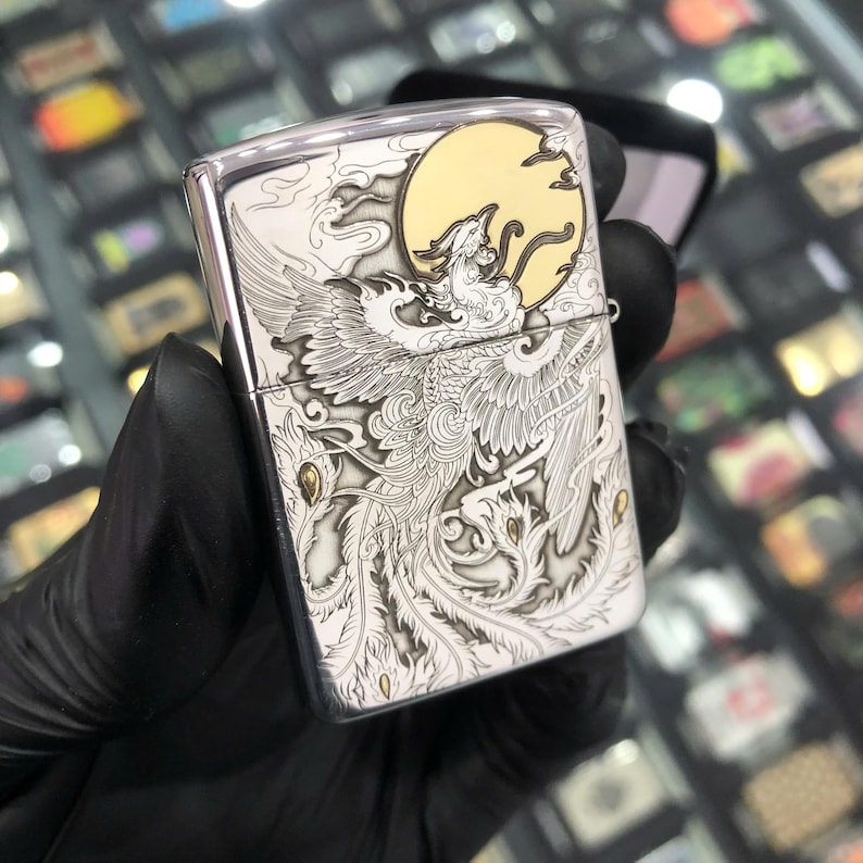 Może przedstawiać: Srebrna metalowa zapalniczka z grawerowanym wzorem feniksa. Zapalniczka posiada szczeg&oacute;łową ilustrację feniksa z pełnią księżyca w tle. Projekt jest w odcieniach srebra i złota.