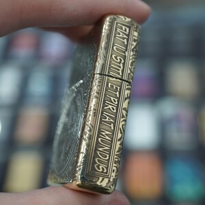 May include: A gold-coloured lighter with detailed engraving and the Latin phrase "Fiat Iustitia Et Pereat Mundus." The rectangular lighter features a hinged lid and ornate patterns. The design has a classic aesthetic.
