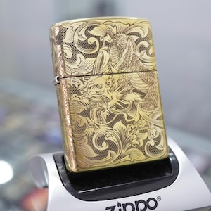 以下が含まれることがあります： 真鍮色のZippoライター。精巧なドラゴンと花の模様が特徴です。詳細な彫刻がライターの表面全体を覆い、クラシックで装飾的なスタイルを示しています。ライターはスタンドに展示されています。