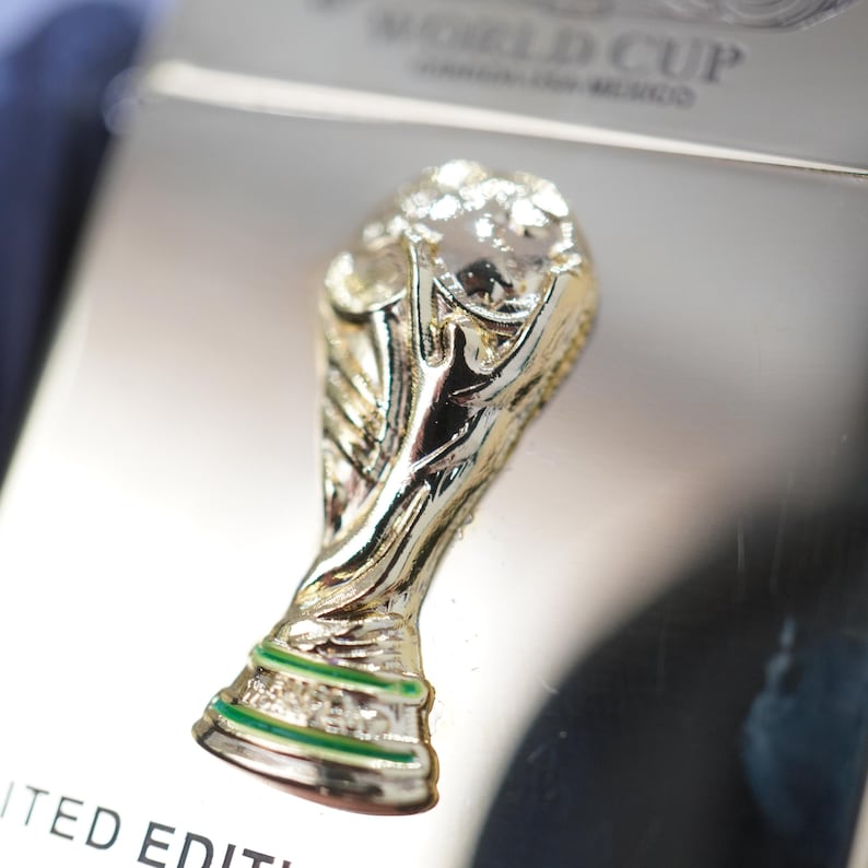 Puede incluir: Un pin de tono dorado con forma de trofeo, que presenta una franja horizontal verde en la base. El pin est&aacute; sujeto a una superficie rectangular de color plateado con el texto WORLD CUP y LIMITED EDITION impreso en letras negras.