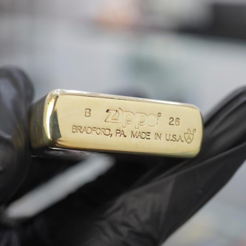 以下が含まれることがあります： B、Zippo、26、およびBRADFORD, PA. MADE IN U.S.A.という刻印と、文字Aが入った小さな盾のロゴが施されたゴールドトーンの金属製Zippoライターの底面。表面は光沢があり、角は丸みを帯びています。