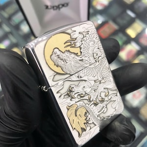 Może przedstawiać: Srebrna zapalniczka Zippo z szczeg&oacute;łowym wzorem smoka. Smok jest wytrawiony w kolorze białym i złotym na srebrnym tle, z ż&oacute;łtym księżycem w tle. Logo Zippo jest widoczne na g&oacute;rze.
