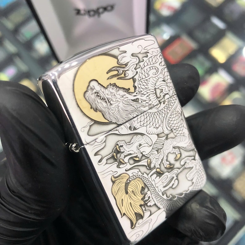 Może przedstawiać: Srebrna zapalniczka Zippo z szczeg&oacute;łowym wzorem smoka. Smok jest wytrawiony w kolorze białym i złotym na srebrnym tle, z ż&oacute;łtym księżycem w tle. Logo Zippo jest widoczne na g&oacute;rze.