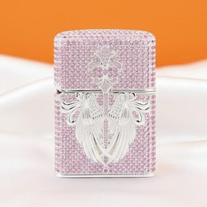 May include: A pink lighter covered in small, sparkling stones. It has a silver-coloured emblem of angel wings and a fleur-de-lis design. The background is a soft orange and white.