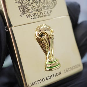 Puede incluir: Un encendedor de metal dorado con un emblema en relieve del trofeo de la Copa del Mundo. La superficie muestra el texto WORLD CUP CANADA USA MEXICO y LIMITED EDITION 1619/2026, junto con un dise&ntilde;o grabado en la parte superior.