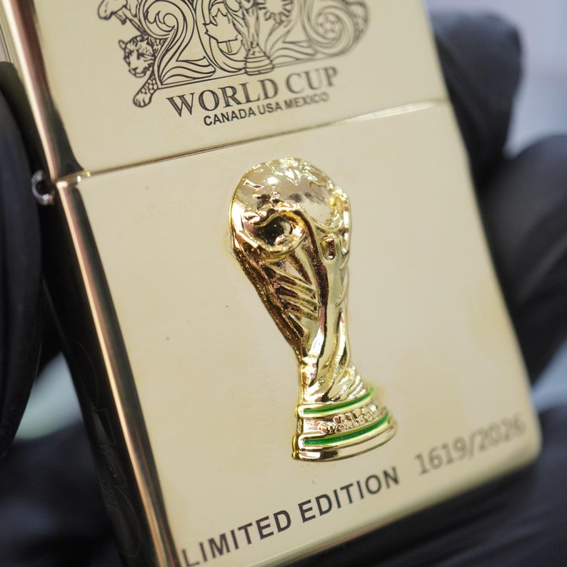 Puede incluir: Un encendedor de metal dorado con un emblema en relieve del trofeo de la Copa del Mundo. La superficie muestra el texto WORLD CUP CANADA USA MEXICO y LIMITED EDITION 1619/2026, junto con un dise&ntilde;o grabado en la parte superior.