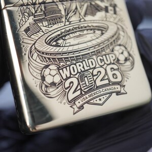 以下が含まれることがあります： 光沢のあるシルバーカラーの金属製ライター。サッカー場、2つのサッカーボール、そして「WORLD CUP 2026 USA-MEXICO-CANADA」という文字が黒いレーザー刻印で詳細に描かれており、数字の0の中には小さなアメリカ国旗のアイコンがあります。
