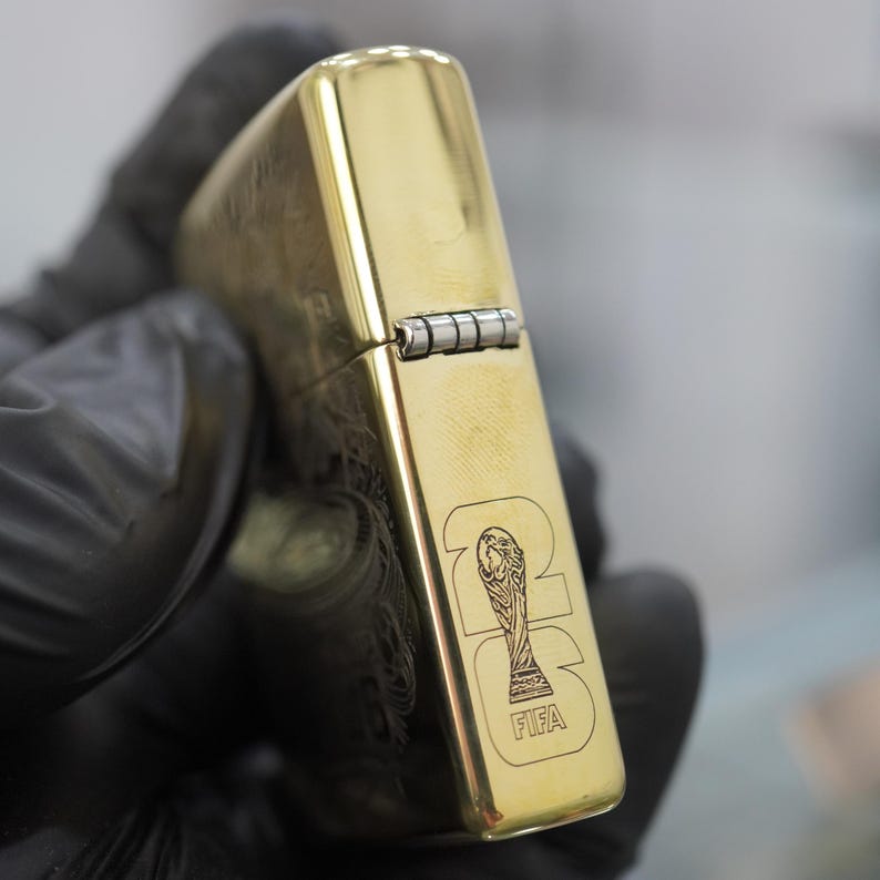 Puede incluir: Un encendedor de metal dorado con tapa abatible, acabado pulido y bisagra plateada. El lateral del encendedor est&aacute; grabado con un gr&aacute;fico del trofeo de la Copa Mundial de la FIFA y el texto 2026, con la palabra FIFA impresa debajo del trofeo.