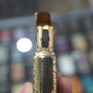 May include: Close-up of a gold-coloured lighter with detailed engraved patterns. The lighter's top is open, showing a black roller and internal parts. The design includes text on the side. A classic accessory.