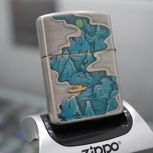 K&ouml;nnte beinhalten: Ein silberfarbenes Zippo-Feuerzeug mit detailliertem Landschaftsdesign. Das Kunstwerk zeigt blaue Berge, ein gelbes Boot und einen gelben Mond vor einem bew&ouml;lkten Himmel. Das Feuerzeug wird in einem silbernen St&auml;nder gehalten, mit dem Zippo-Logo.
