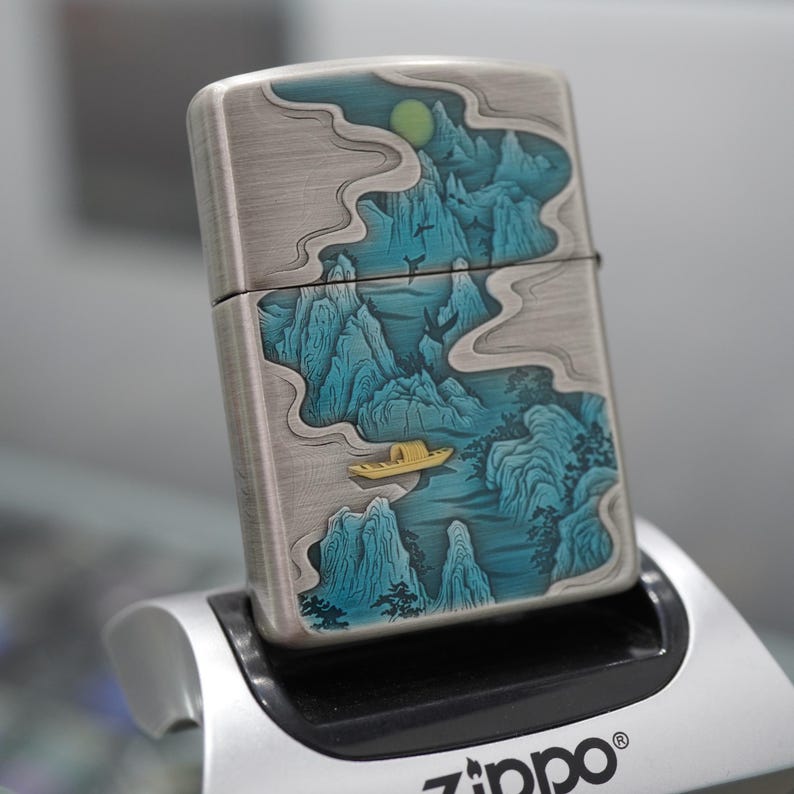 K&ouml;nnte beinhalten: Ein silberfarbenes Zippo-Feuerzeug mit detailliertem Landschaftsdesign. Das Kunstwerk zeigt blaue Berge, ein gelbes Boot und einen gelben Mond vor einem bew&ouml;lkten Himmel. Das Feuerzeug wird in einem silbernen St&auml;nder gehalten, mit dem Zippo-Logo.