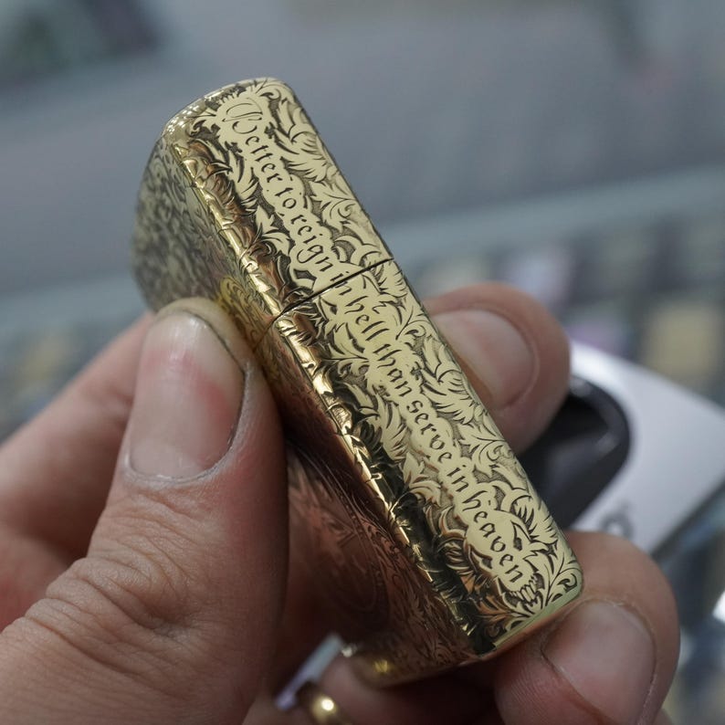 May include: A gold-coloured lighter with detailed floral engravings. The lighter has the quote "Better to reign in hell than serve in heaven" in a decorative font. The lighter is held in a person's hand.