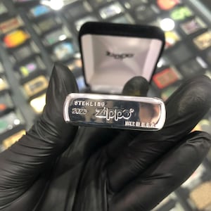 Może przedstawiać: Srebrna zapalniczka Zippo z napisem "STERLING 2023 Zippo MADE IN U.S.A." jest wyświetlana. Zapalniczka znajduje się w czarnym aksamitnym pudełku. Zapalniczka jest trzymana przez osobę w czarnych rękawicach.