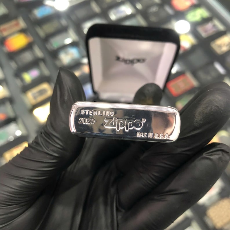 Może przedstawiać: Srebrna zapalniczka Zippo z napisem "STERLING 2023 Zippo MADE IN U.S.A." jest wyświetlana. Zapalniczka znajduje się w czarnym aksamitnym pudełku. Zapalniczka jest trzymana przez osobę w czarnych rękawicach.