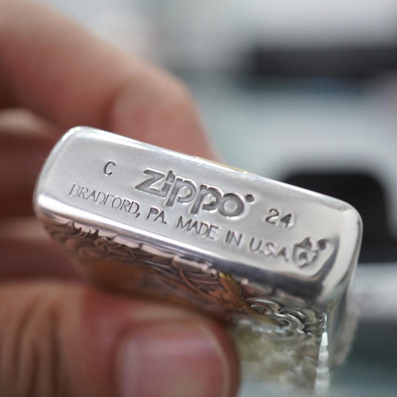 Może przedstawiać: Zbliżenie srebrnej zapalniczki Zippo. Zapalniczka ma skomplikowane grawerowane wzory, a tekst "Zippo" i "BRADFORD, PA. MADE IN U.S.A." jest widoczny. Zapalniczka jest trzymana w dłoni.