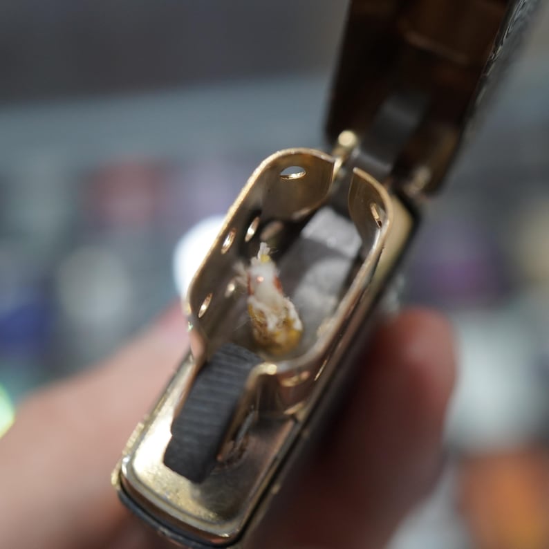 May include: An open, gold-coloured metal lighter, showcasing its internal components. The lighter contains a wick and cotton. The design is vintage-inspired, with the lighter held open to display its features.