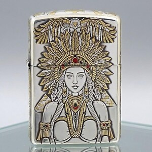 May include: A rectangular metal lighter featuring an intricate embossed design of a person wearing a large feathered headdress topped with an eagle. The artwork includes gold and silver tones with small red and green accents and two pyramids.