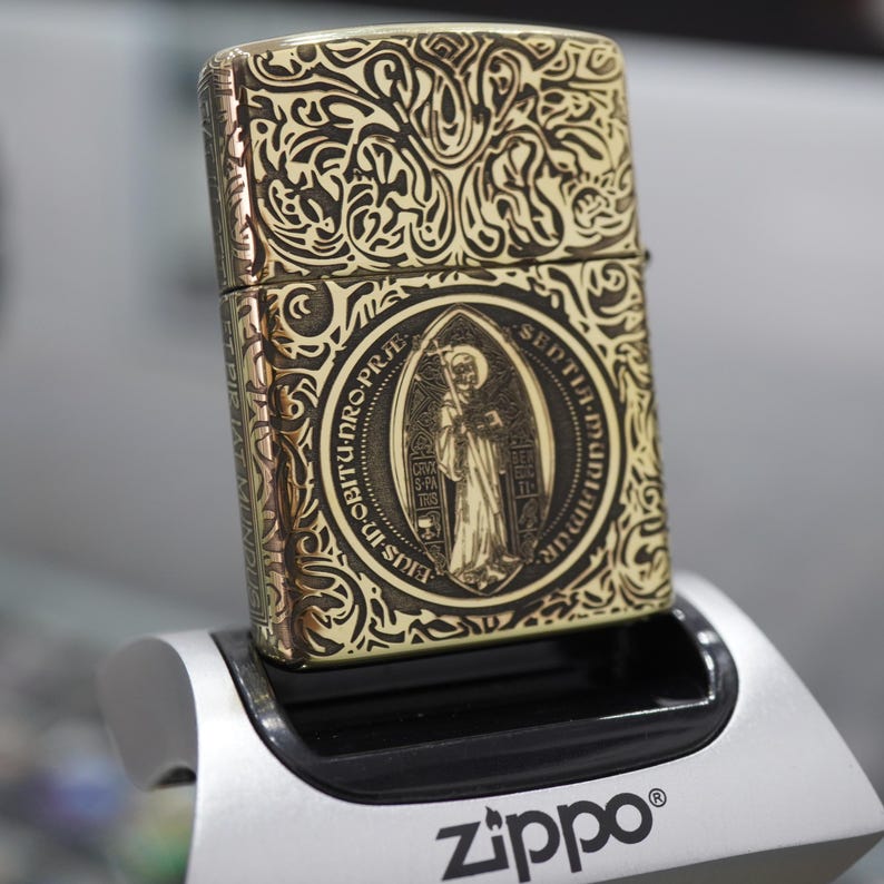 May include: A gold-coloured Zippo lighter with detailed engraved designs and a central medallion depicting a religious figure. The lighter is on a silver and black stand, with the Zippo logo visible.