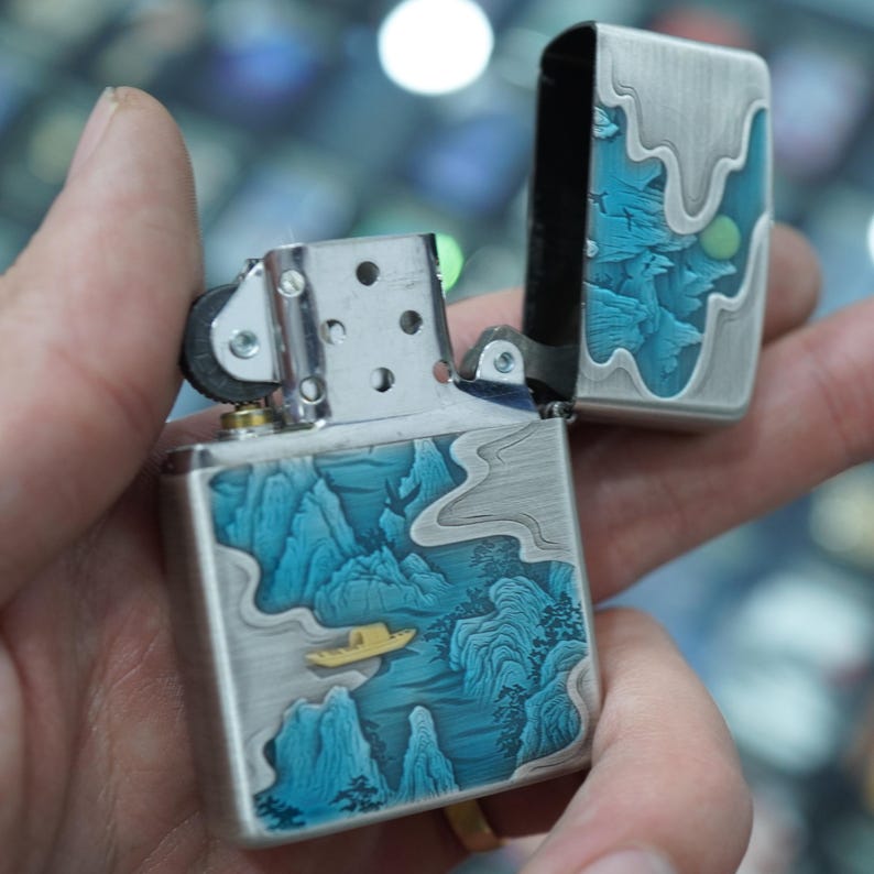 K&ouml;nnte beinhalten: Ein silberfarbenes Feuerzeug mit offenem Deckel, das ein detailliertes blau-graues Landschaftsdesign aufweist. Das Design umfasst Berge, Wasser und ein kleines gelbes Boot. Das Feuerzeug wird in einer Hand offen gehalten.