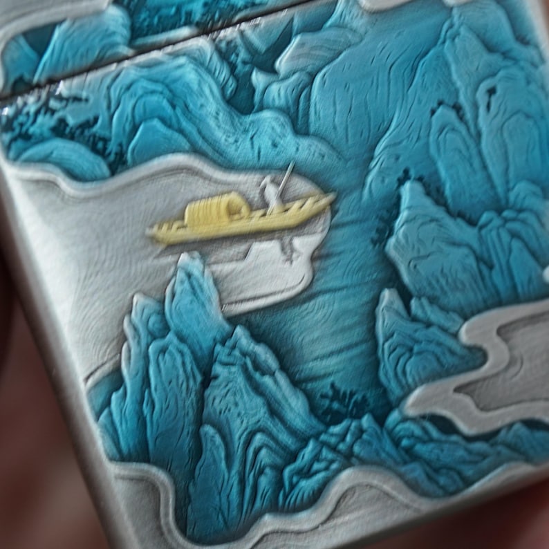 K&ouml;nnte beinhalten: Ein silberfarbenes Feuerzeug mit einem detaillierten Reliefdesign, das eine Landschaft darstellt. Das Design umfasst blaue Berge, ein gelbes Boot mit einer Figur und eine Flussszene. Die Oberfl&auml;che des Feuerzeugs hat eine strukturierte Optik.
