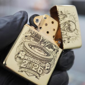 以下が含まれることがあります： ゴールドトーンの金属製フリップトップライター。スタジアム、サッカーボール、「WORLD CUP 2026 USA-MEXICO-CANADA」の文字が黒いレーザー刻印で施されています。蓋が開いており、内部の真鍮色のチムニーとフリントホイールが見えています。
