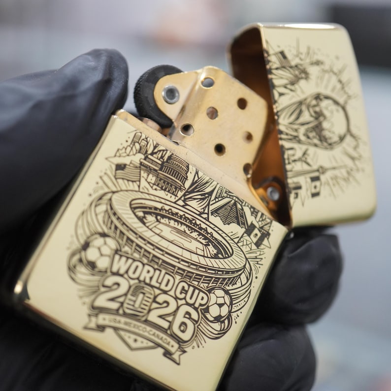 以下が含まれることがあります： ゴールドトーンの金属製フリップトップライター。スタジアム、サッカーボール、「WORLD CUP 2026 USA-MEXICO-CANADA」の文字が黒いレーザー刻印で施されています。蓋が開いており、内部の真鍮色のチムニーとフリントホイールが見えています。