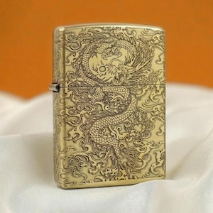 Pode incluir: Um isqueiro de metal dourado com um desenho de drag&atilde;o gravado. O drag&atilde;o &eacute; detalhado, rodeado por nuvens rodopiantes. O isqueiro tem formato retangular com tampa articulada e dobradi&ccedil;a prateada.