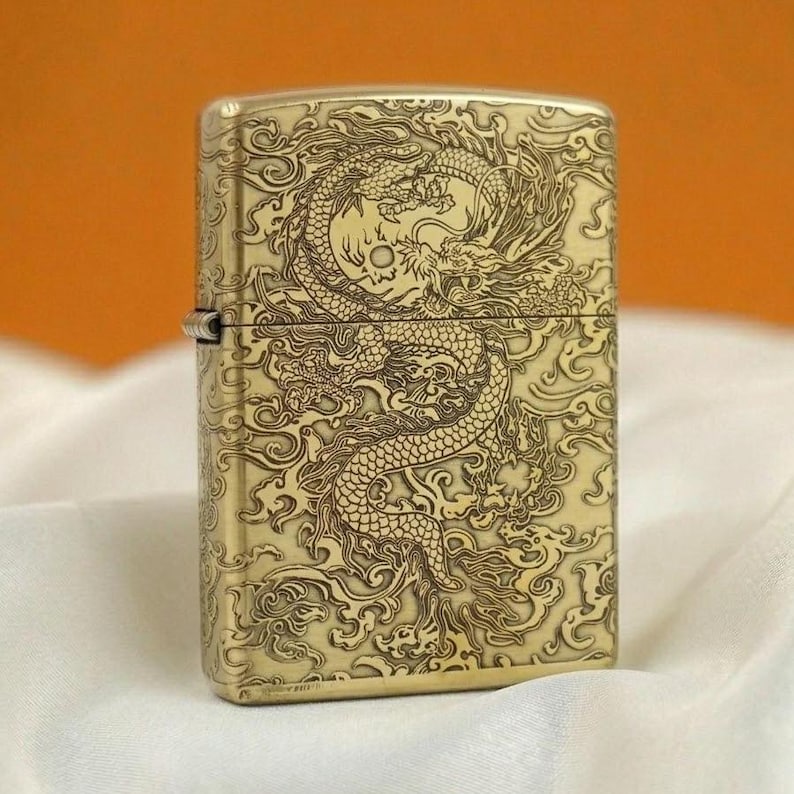 Pode incluir: Um isqueiro de metal dourado com um desenho de drag&atilde;o gravado. O drag&atilde;o &eacute; detalhado, rodeado por nuvens rodopiantes. O isqueiro tem formato retangular com tampa articulada e dobradi&ccedil;a prateada.