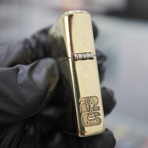 以下が含まれることがあります： 側面に長方形のヒンジが付いた、光沢のあるゴールドトーンの金属製フリップトップライター。表面にはFIFAワールドカップトロフィーのグラフィックと、ライターの前面下部にFIFAの文字が刻印されています。
