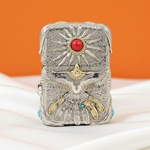 K&ouml;nnte beinhalten: Silberfarbenes Metallfeuerzeug mit aufwendigen gepr&auml;gten Designs. Es hat einen roten Stein oben und ein Adler-Motiv mit goldfarbenen Akzenten. Kleine t&uuml;rkisfarbene Steine sind an den Seiten eingesetzt.