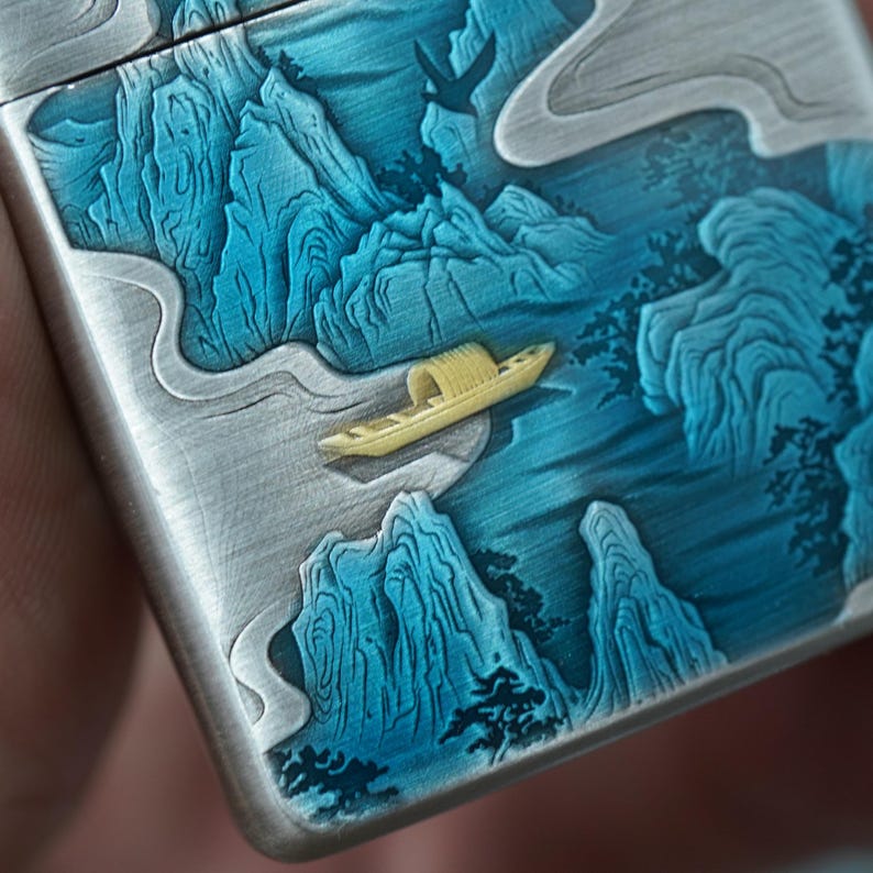 K&ouml;nnte beinhalten: Ein silberfarbenes Metallfeuerzeug mit einem detaillierten Landschaftsdesign. Das Design zeigt blaue Berge, ein gelbes Boot und einen fliegenden Vogel. Das Kunstwerk ist aufwendig geschnitzt und bemalt und erzeugt einen dreidimensionalen Effekt.