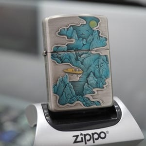K&ouml;nnte beinhalten: Silbernes Zippo-Feuerzeug mit detailliertem Landschaftsdesign mit t&uuml;rkisfarbenen Bergen, einem gelben Boot und einem Vollmond. Das Feuerzeug wird auf einem silbernen St&auml;nder mit dem Zippo-Logo pr&auml;sentiert.