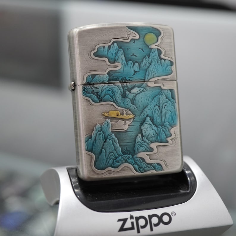 K&ouml;nnte beinhalten: Silbernes Zippo-Feuerzeug mit detailliertem Landschaftsdesign mit t&uuml;rkisfarbenen Bergen, einem gelben Boot und einem Vollmond. Das Feuerzeug wird auf einem silbernen St&auml;nder mit dem Zippo-Logo pr&auml;sentiert.