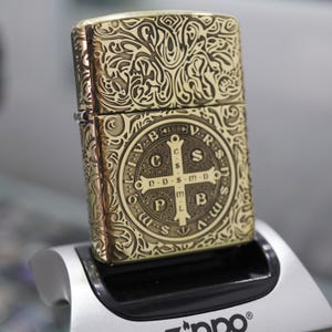 May include: A gold-coloured Zippo lighter with intricate engraved patterns and a cross design. The lighter features a detailed, ornate design on its surface, with a classic rectangular shape. The lighter is displayed on a silver stand.