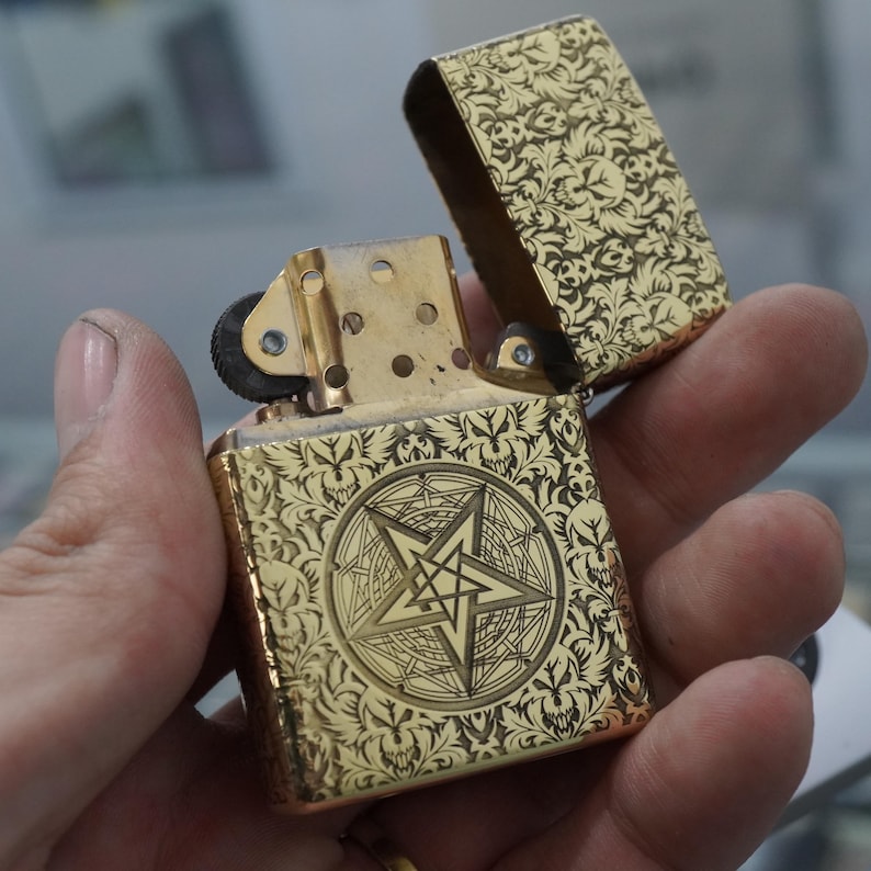 May include: A gold-coloured lighter with an open lid, held in a hand. The lighter features an engraved floral pattern and a central pentagram design. The lighter's surface has a shiny, metallic finish.