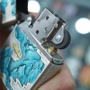 K&ouml;nnte beinhalten: Ein silberfarbenes Feuerzeug mit einem detaillierten blau-wei&szlig;en Wellendesign. Die Oberseite des Feuerzeugs ist ge&ouml;ffnet und zeigt den Z&uuml;ndmechanismus. Das Design zeigt ein kleines gelbes Boot und f&uuml;gt ein maritimes Thema hinzu.