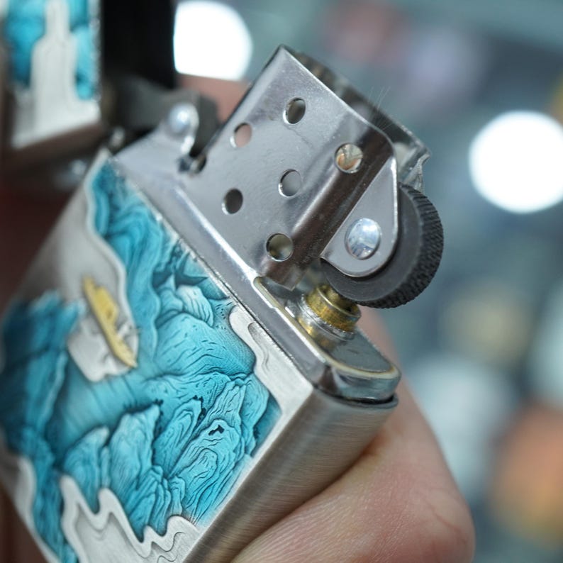 K&ouml;nnte beinhalten: Ein silberfarbenes Feuerzeug mit einem detaillierten blau-wei&szlig;en Wellendesign. Die Oberseite des Feuerzeugs ist ge&ouml;ffnet und zeigt den Z&uuml;ndmechanismus. Das Design zeigt ein kleines gelbes Boot und f&uuml;gt ein maritimes Thema hinzu.