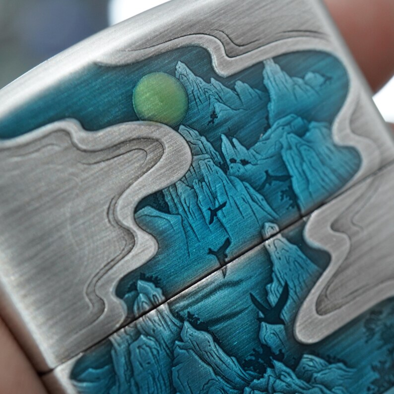 K&ouml;nnte beinhalten: Ein silberfarbenes Metallfeuerzeug mit detailliertem Landschaftsdesign. Das Design zeigt blaue Berge, einen gr&uuml;nen Mond und fliegende V&ouml;gel. Das Feuerzeug hat eine geb&uuml;rstete Metalloberfl&auml;che und einen Klappdeckel.