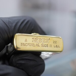 Puede incluir: Una base de encendedor Zippo de metal dorado con texto grabado que dice A, Zippo, 22 y BRADFORD, PA. MADE IN U.S.A. La superficie inferior rectangular presenta un acabado met&aacute;lico pulido con letras estampadas claras y una peque&ntilde;a bisagra.