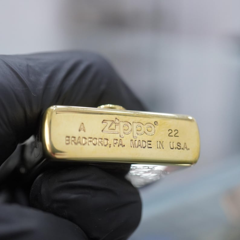 Puede incluir: Una base de encendedor Zippo de metal dorado con texto grabado que dice A, Zippo, 22 y BRADFORD, PA. MADE IN U.S.A. La superficie inferior rectangular presenta un acabado met&aacute;lico pulido con letras estampadas claras y una peque&ntilde;a bisagra.