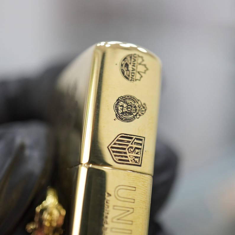 Puede incluir: Un encendedor de metal dorado pulido con tres emblemas grabados distintos en el lateral. El emblema superior representa una hoja de arce, el central muestra un escudo circular y el inferior es un escudo con las letras USF. La palabra UNITED es visible.