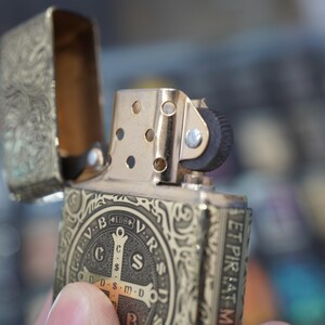 May include: Gold-coloured metal lighter with an open lid, featuring intricate engraved patterns and a religious emblem. The lighter's design includes a cross and the text "V.R.S.N.S.M.D."