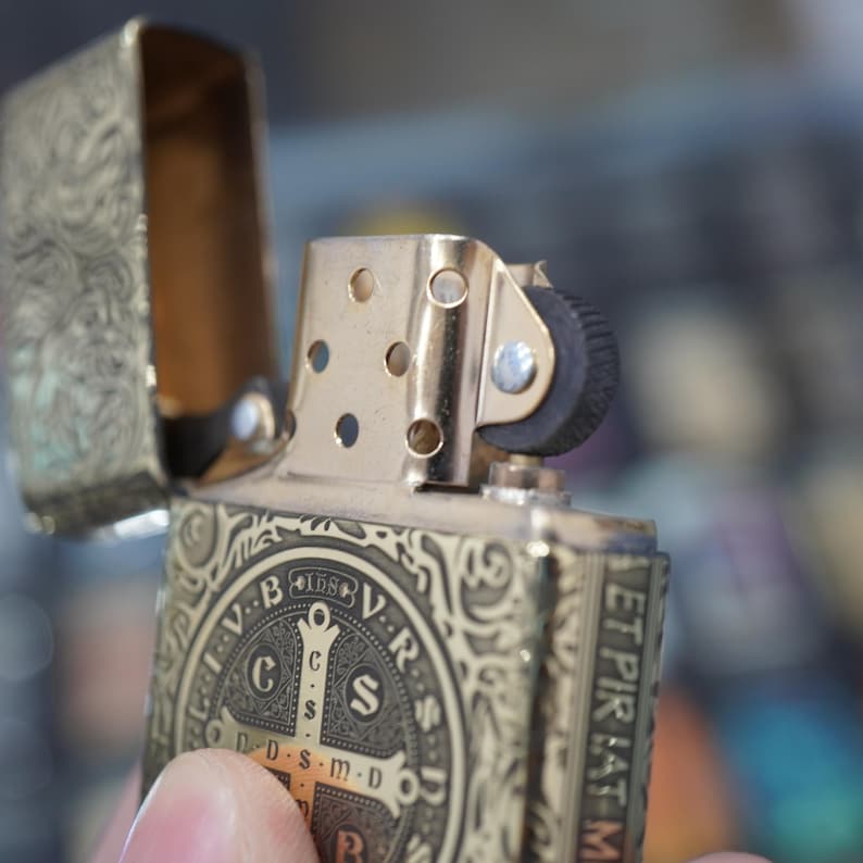 May include: Gold-coloured metal lighter with an open lid, featuring intricate engraved patterns and a religious emblem. The lighter's design includes a cross and the text "V.R.S.N.S.M.D."