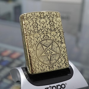May include: Gold-coloured metal lighter with an ornate pattern of skulls and floral designs. The front features a pentagram within a circle. The lighter is resting on a black and silver display stand.