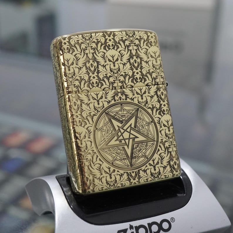 May include: Gold-coloured metal lighter with an ornate pattern of skulls and floral designs. The front features a pentagram within a circle. The lighter is resting on a black and silver display stand.