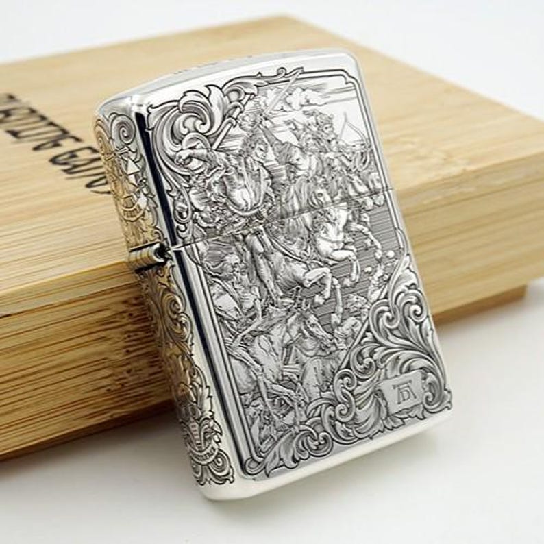 May include: Silver-colored metal lighter with an intricate engraved design depicting a battle scene with figures on horseback. The lighter is rectangular and has ornate scrollwork around the edges. It rests on a wooden box.