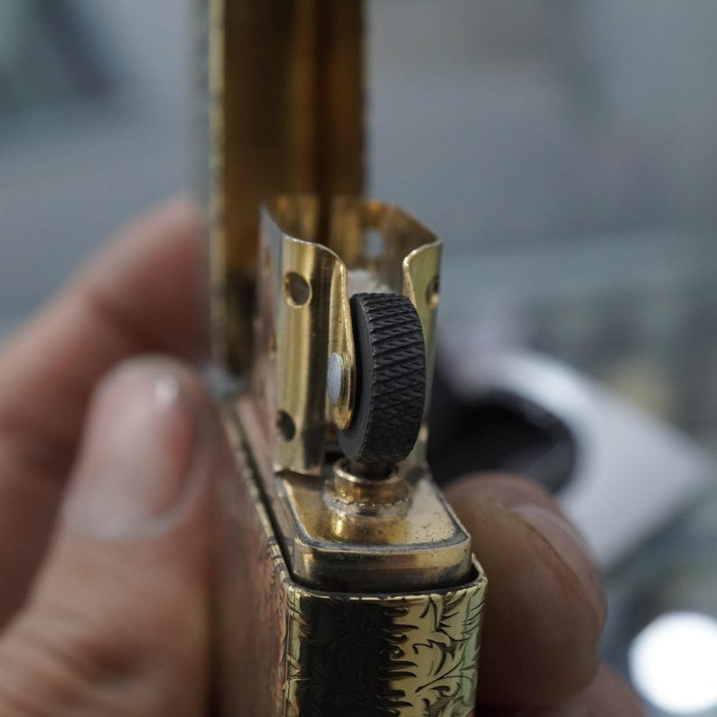 May include: Close-up of a gold-coloured lighter with a black textured roller. The lighter is open, revealing its internal components. The body of the lighter features an engraved floral pattern.