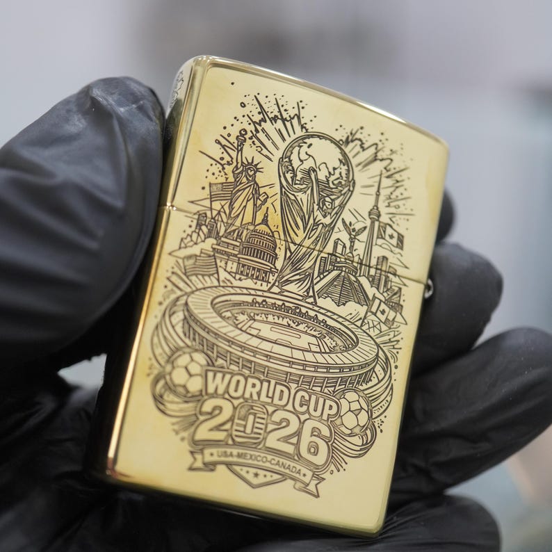 Puede incluir: Un encendedor de metal dorado con un grabado l&aacute;ser negro detallado del trofeo de la Copa Mundial 2026, un estadio y monumentos ic&oacute;nicos de EE. UU., M&eacute;xico y Canad&aacute;, con el texto "WORLD CUP 2026 USA-MEXICO-CANADA" claramente visible.