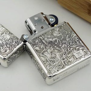 May include: A silver-colored lighter with intricate engraved designs. The lighter is open, revealing the interior components. The detailed carvings depict figures and ornate patterns. The lighter is marked "STERLING 2012".