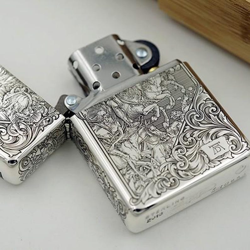 May include: A silver-colored lighter with intricate engraved designs. The lighter is open, revealing the interior components. The detailed carvings depict figures and ornate patterns. The lighter is marked "STERLING 2012".